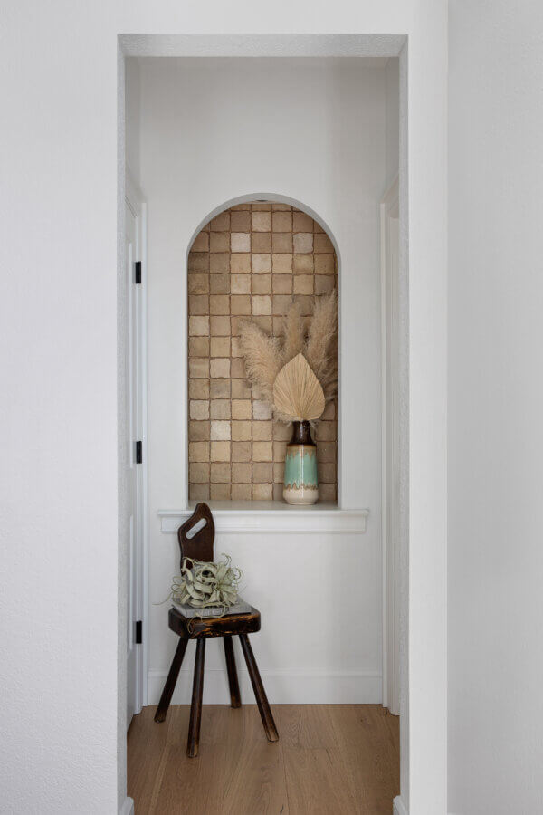 Step Inside an Austin Interior Designer’s Home - 6 A small alcove with tan square tiles creates a personal retreat, displaying a ceramic vase with dried palm leaves and pampas grass. A wooden stool with a decorative object sits on the light wood floor below.