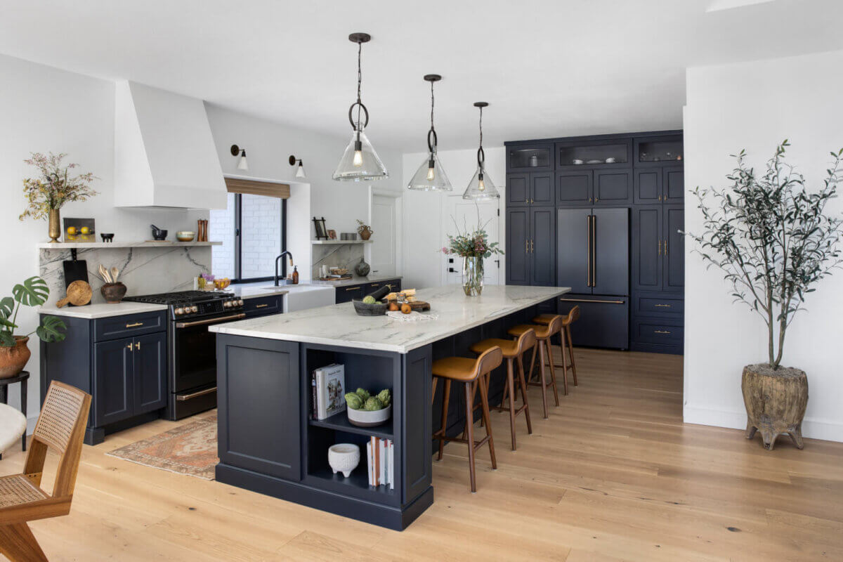 Modern kitchen by an Austin interior designer featuring navy cabinets, a large marble island with four brown stools, pendant lights, wood flooring, and decorative plants—a true personal retreat.