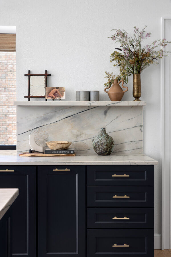 Step Inside an Austin Interior Designer’s Home - 9 A modern kitchen corner designed by an Austin interior designer features dark cabinets, a marble backsplash, books, pottery, and a vase with wildflowers arranged on a shelf, creating a perfect personal retreat.