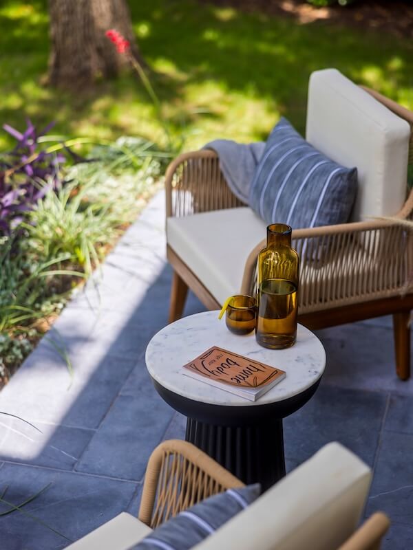 Step Inside an Austin Interior Designer’s Home - 17 Outdoor patio scene with two cushioned chairs, a small round table holding amber glassware, and a book—an inviting personal retreat set on gray stone tiles surrounded by green plants.