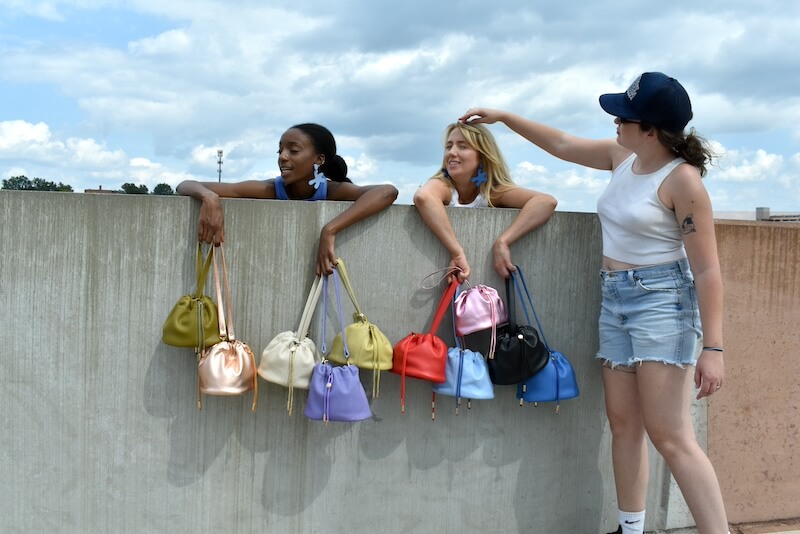 Cold Gold Standard: Bold Leather Bags & Jewelry - 2 Three women on a rooftop, two leaning over a concrete wall displaying colorful handbags, while a third woman in a hat playfully reaches toward one of the cold gold accessories.