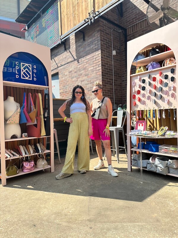 Cold Gold Standard: Bold Leather Bags & Jewelry - 4 Two women stand in front of a market stall displaying cold gold handbags, jewelry, and accessories, with brick walls and outdoor sunlight in the background.