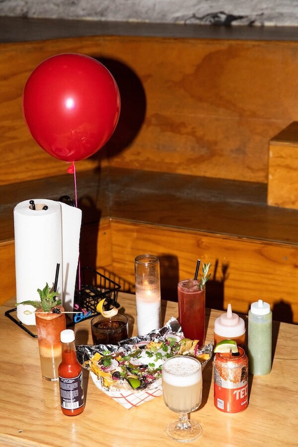 20 Epic Spots to Grab Dinner at the Bar in Nashville - 4 A wooden table set for dinner at the bar, featuring assorted cocktails, nachos, condiments, a roll of paper towels, candles, and a red balloon attached to the holder.