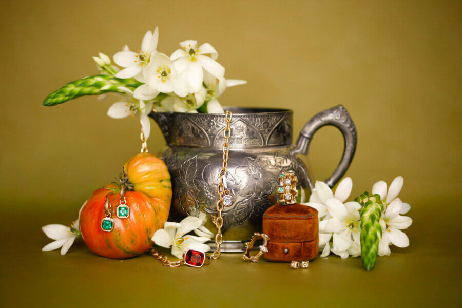 Heirloom Jewelry, Reimagined: Meet Designer Brooke Griffith - 4 A silver pitcher with white flowers, a striped orange tomato, heirloom jewelry, and a small brown box are arranged against an olive background.