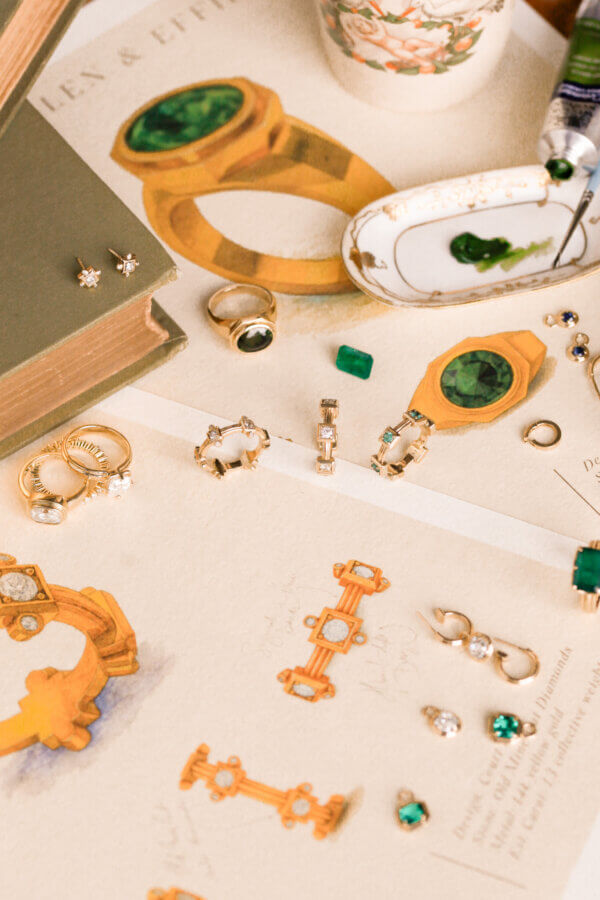 Heirloom Jewelry, Reimagined: Meet Designer Brooke Griffith - 5 Assorted heirloom jewelry with green and white gemstones arranged on sketch papers with ring designs, next to a book, a floral cup, and a paint tube—capturing the creative spirit of a Nashville artist.