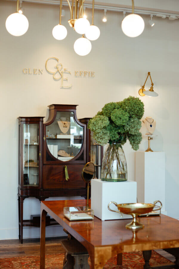 Heirloom Jewelry, Reimagined: Meet Designer Brooke Griffith - 3 A chic jewelry boutique interior featuring wooden furniture, heirloom jewelry in a display cabinet, a vase with green hydrangeas, and a gold logo on the wall reading "Glen Effie," showcasing the work of a Nashville artist.