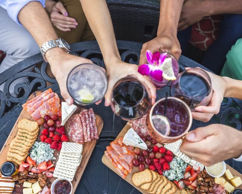 Five people toast with drinks over a table set with charcuterie boards featuring crackers, cheese, fruit, and cured meats—capturing the relaxed spirit of beach vacations.