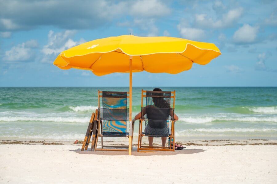 A person enjoys beach vacations, sitting on a wooden chair under a large yellow umbrella facing the ocean, with another empty chair beside them on the sandy shore.