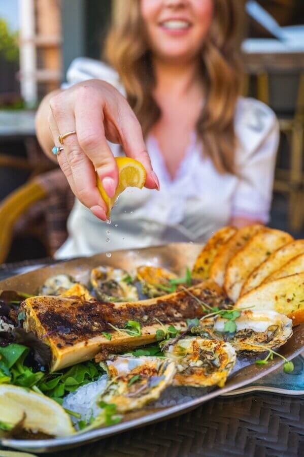A woman squeezes a lemon wedge over a platter of grilled bone marrow, oysters, and toasted bread slices on a table, evoking the flavors of unforgettable beach vacations.