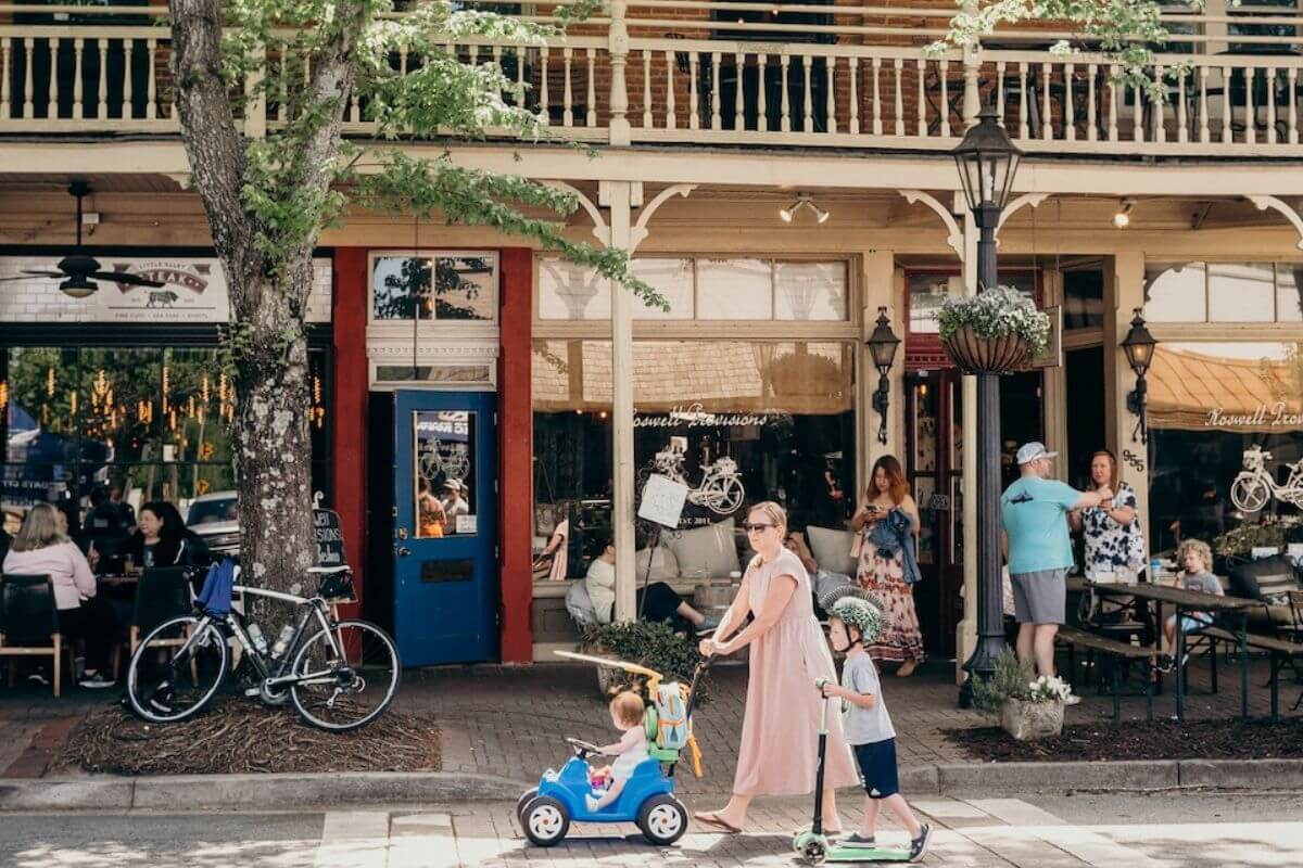 A woman walks with two children on scooters past shops and outdoor diners, enjoying a sunny day in a small downtown area—it's the perfect way to shop in Georgia.
