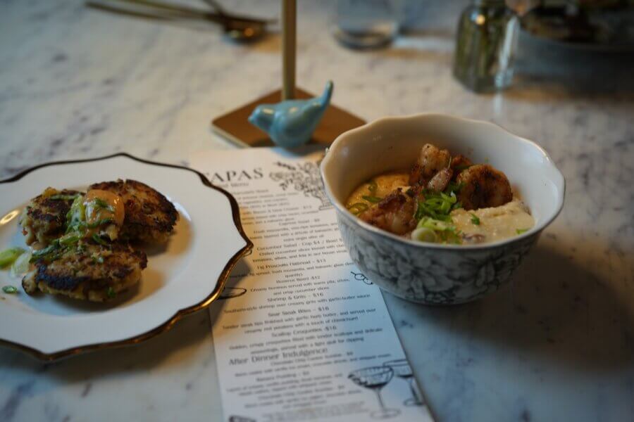 How to Eat, Shop & Explore Your Way Through Georgia - 3 A bowl of shrimp and grits and a plate with two crab cakes sit next to a printed tapas menu on a marble table—perfect for those looking to Eat in Georgia.