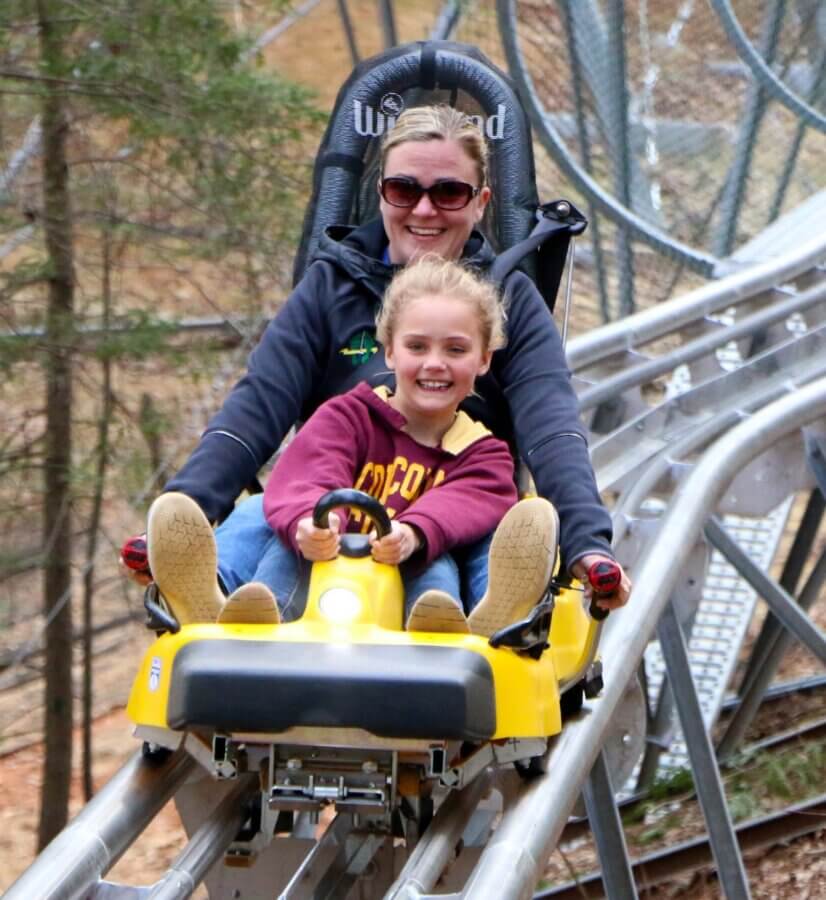 How to Eat, Shop & Explore Your Way Through Georgia - 5 An adult and a child ride a yellow roller coaster together, both smiling, on a track through a wooded area—just one exciting way to play in Georgia.