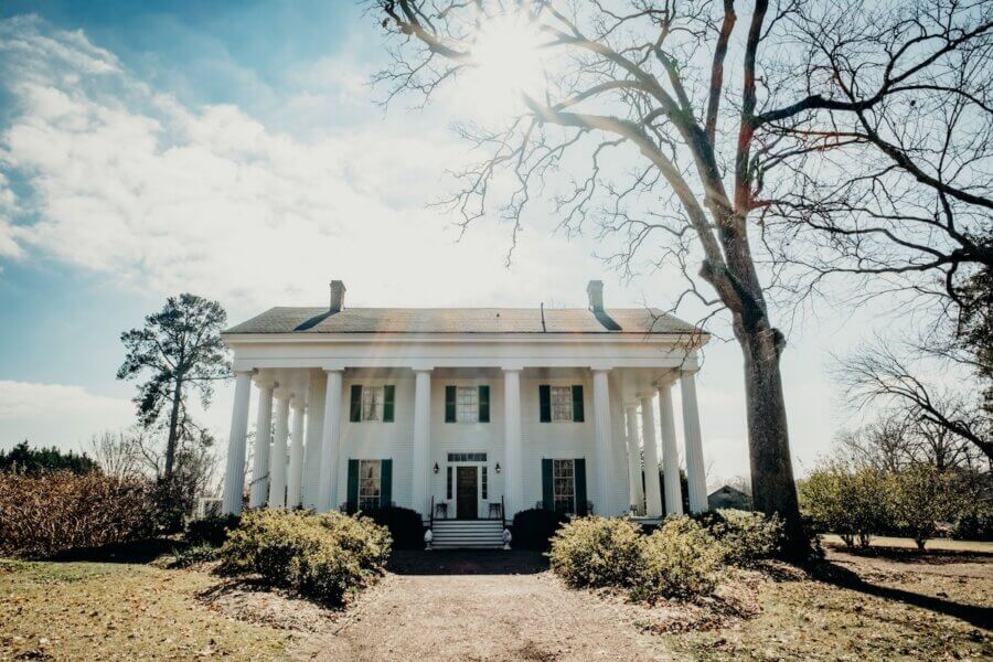 How to Eat, Shop & Explore Your Way Through Georgia - 6 A large white two-story house with tall columns and a front porch sits under a bright sun and clear sky, surrounded by bushes and leafless trees—perfect for those looking to explore, shop in Georgia, or simply enjoy southern charm.