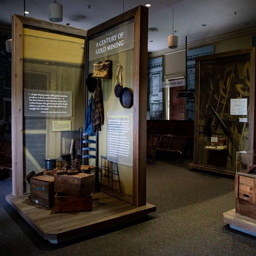How to Eat, Shop & Explore Your Way Through Georgia - 12 A museum exhibit titled “A Century of Gold Mining” displays mining tools, boots, hats, and historical information panels in a dimly lit room—perfect for those looking to dig into history while they Eat Georgia, Play Georgia, and Shop Georgia.