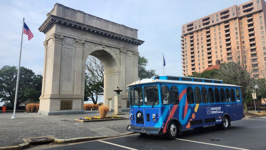 9 Hidden Gems to Visit in the South - 10 A blue Newport News Tourist trolley is parked in front of the grand stone "Victory" arch, with an American flag and tall building behind.