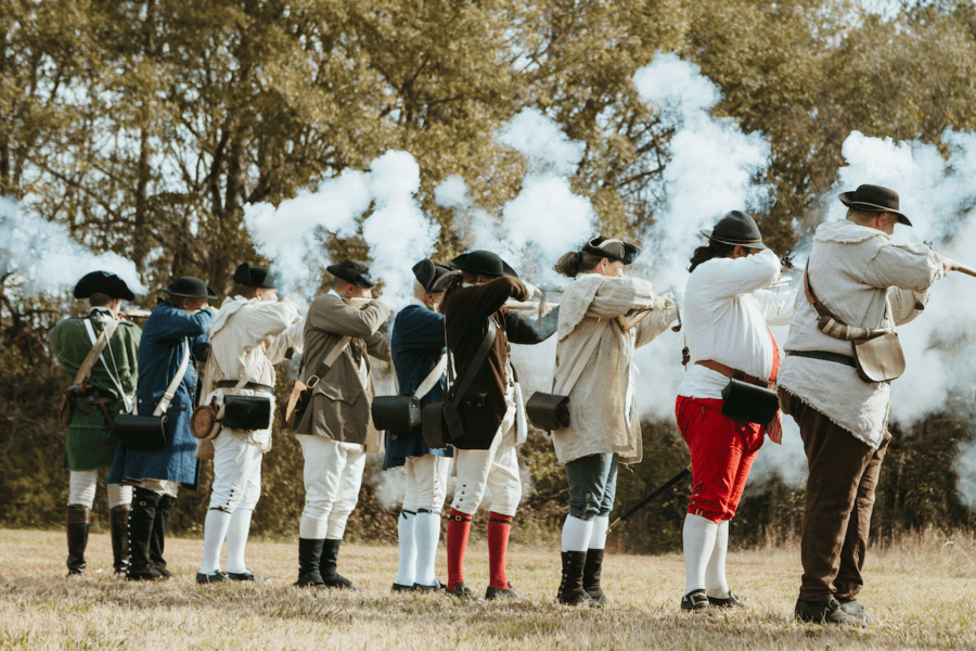 9 Hidden Gems to Visit in the South - 12 A group of people in historical clothing fire muskets outdoors, reenacting a battle scene in the South, with smoke visible in the air—one of the region’s hidden gems for history enthusiasts.