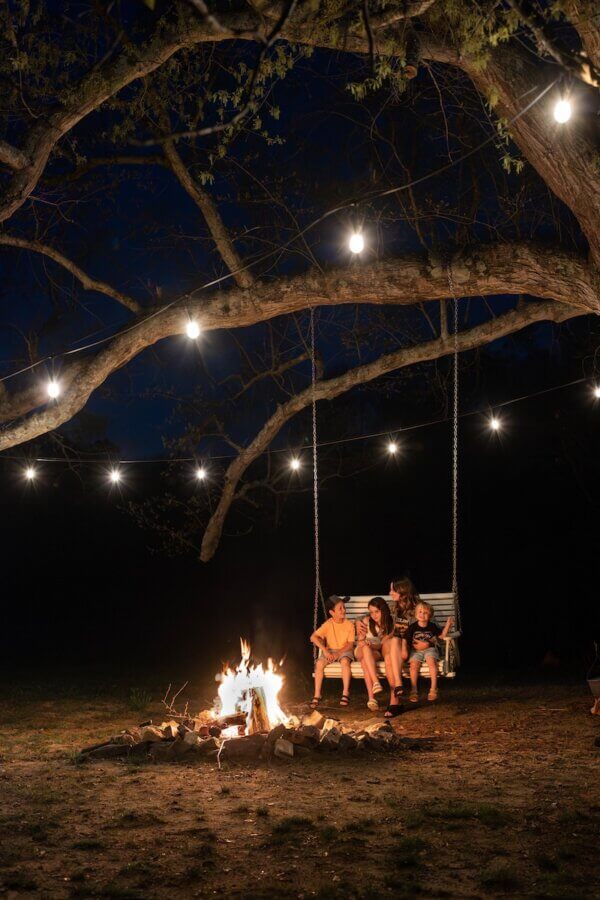 9 Hidden Gems to Visit in the South - 13 Four people sit on a swing under string lights near a campfire at night, surrounded by trees—a true hidden gem in the South.