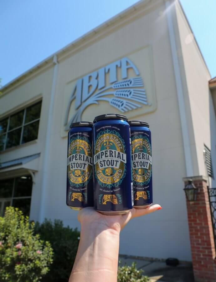 Savoring Louisiana: A Culinary Roadshow Across the State - 9 A hand holds up four cans of Abita Imperial Stout in front of a building with a large Abita sign, capturing the spirit of Louisiana.