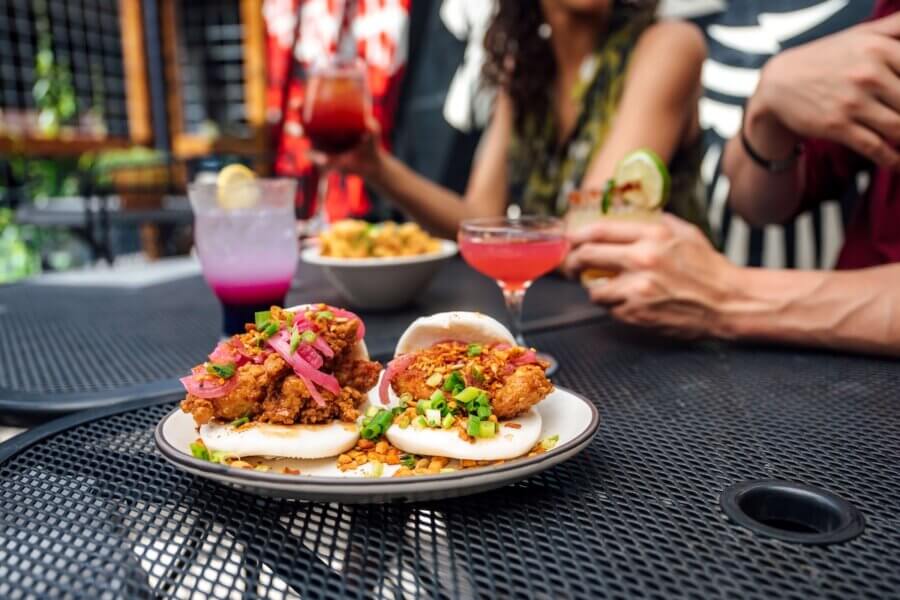 Savoring Louisiana: A Culinary Roadshow Across the State - 3 Two plates of bao buns with fried filling and garnish sit on an outdoor table in Louisiana, while three people hold drinks in the background.
