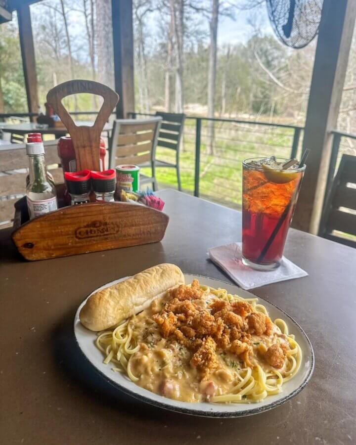Savoring Louisiana: A Culinary Roadshow Across the State - 8 Plate of pasta with creamy sauce and fried chicken, served with a breadstick, sits next to a glass of iced tea on an outdoor table overlooking a wooded Louisiana landscape.