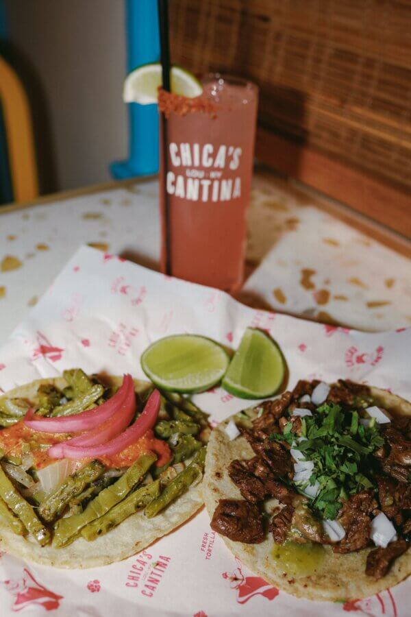 4 New Louisville Restaurants (+2 New Menus!) to Try This Month - 8 Two tacos, one with grilled cactus, onions, and pickled onions, and one with beef, onions, and cilantro, are served with lime wedges next to a pink drink in a glass labeled "Chica's Cantina.