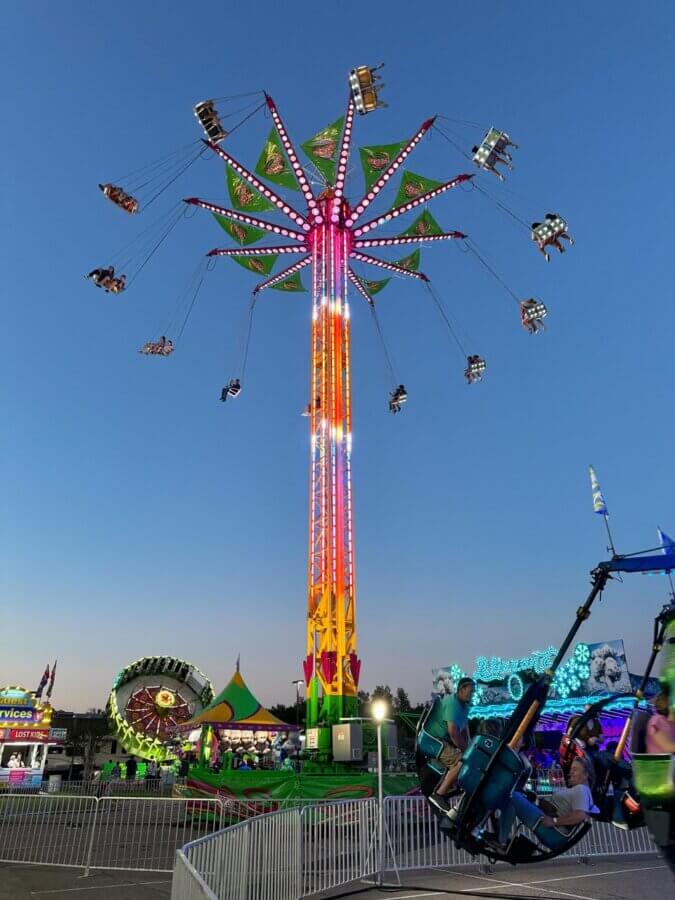 6 Mississippi Spring Music Festivals Not to Miss - 2 A tall illuminated swing ride spins people in the air at an outdoor fair during dusk, with other carnival rides and booths visible in the background.