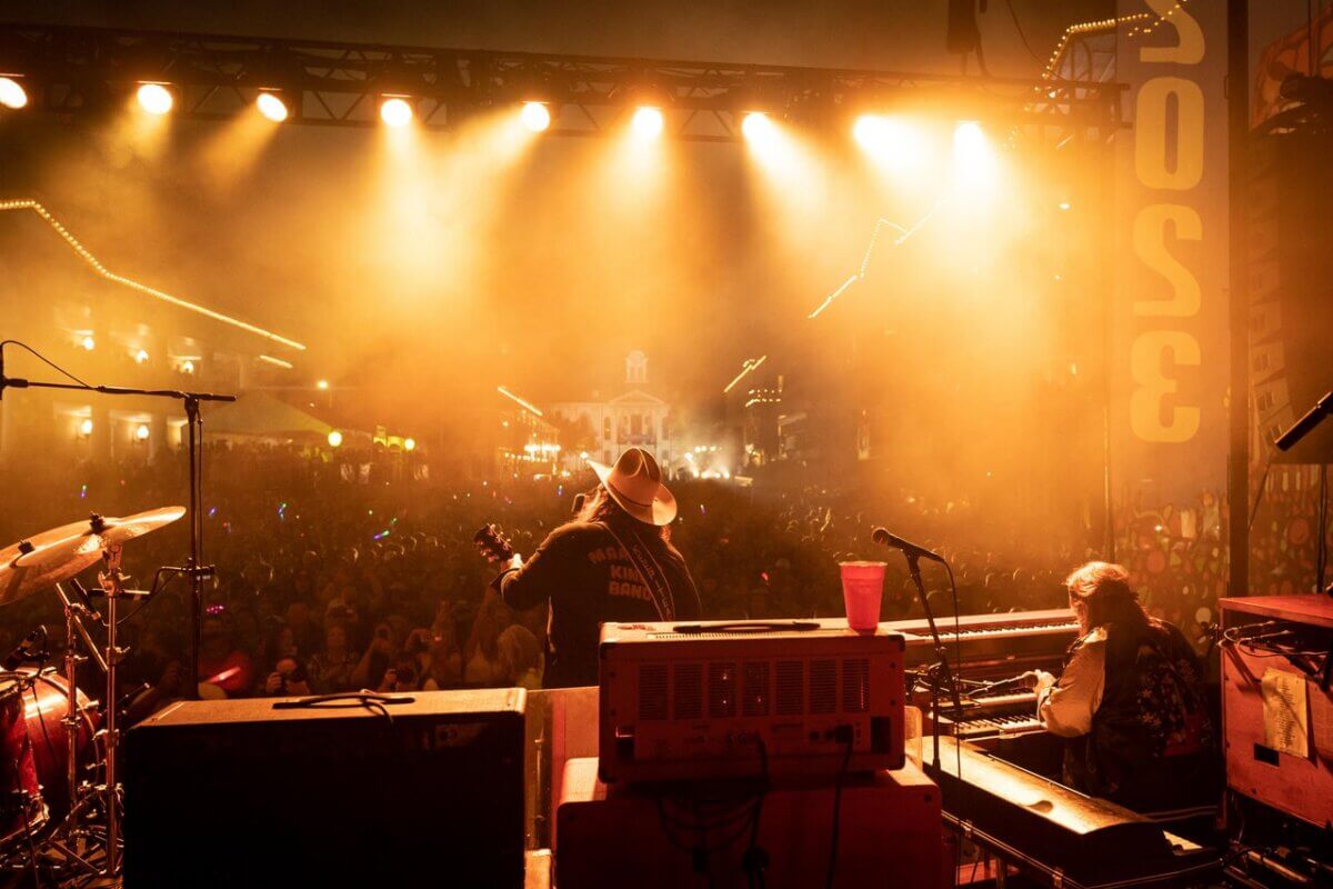 Musicians perform on a stage under bright lights at night, facing a large crowd at an outdoor event—capturing the vibrant energy of spring music festivals in Mississippi.
