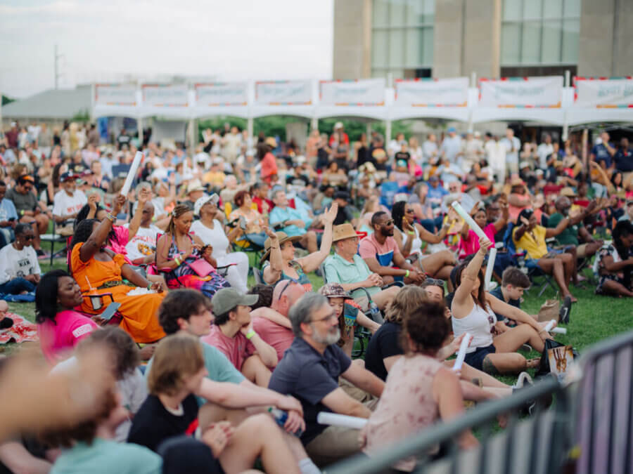 6 Mississippi Spring Music Festivals Not to Miss - 5 A large crowd of people sits on the grass at an outdoor event, some raising their hands, with banners and a building in the background—capturing the lively spirit of Mississippi spring festivals.
