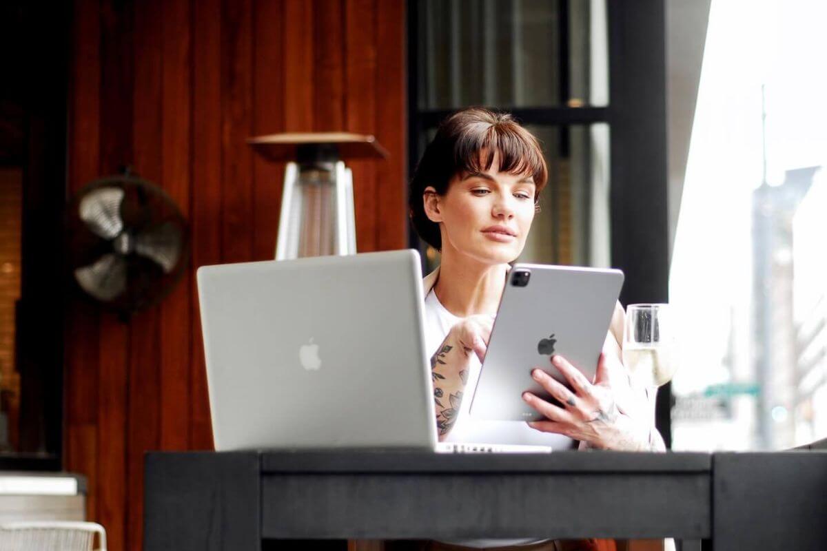 Nicolette Tomblyn sits at a table with a laptop and tablet, focusing on the tablet screen. A glass of water rests nearby, while the wooden wall in the background adds warmth to this FACES-inspired scene.