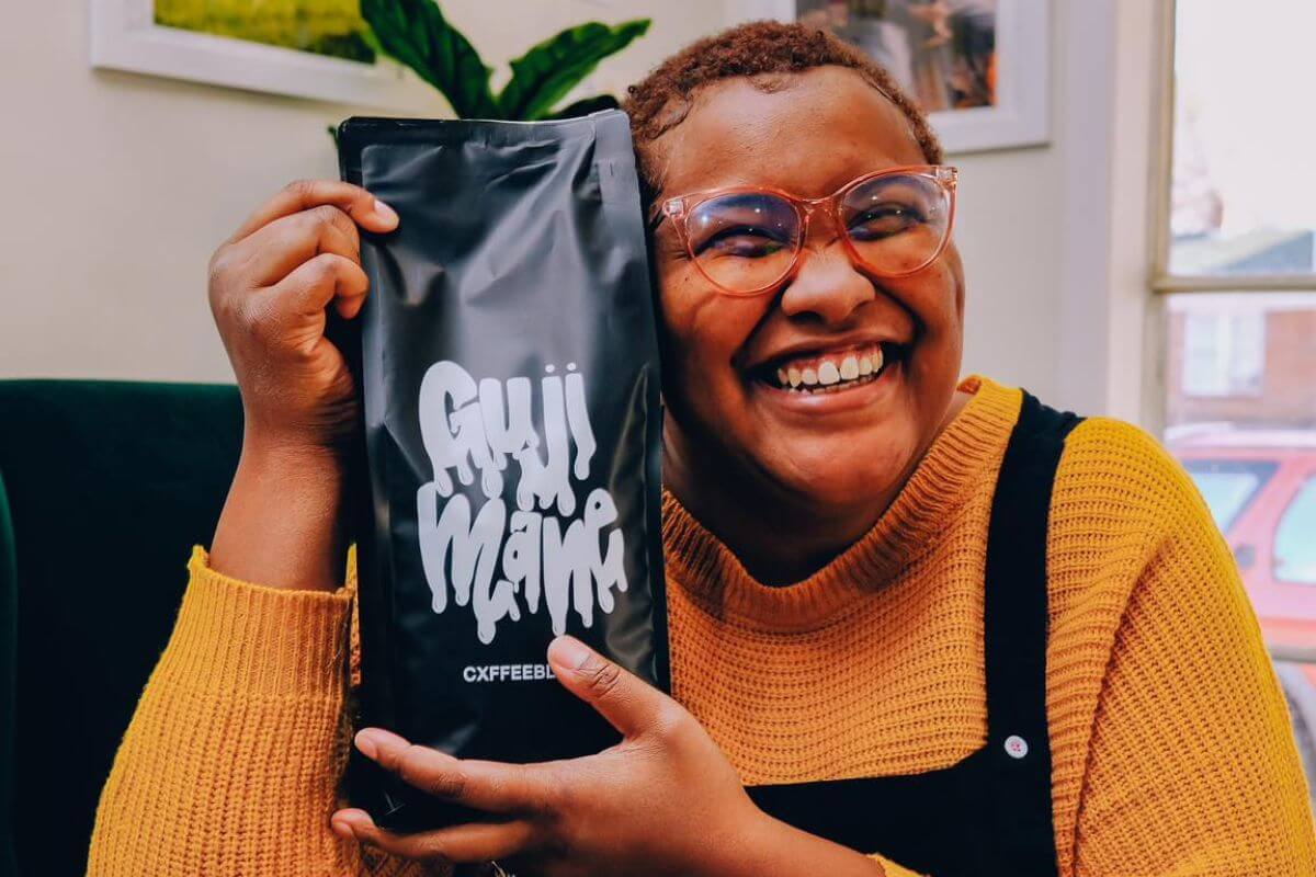 Renata Henderson smiles while holding up a black "Guji Mama CXFFEEBLACK" bag, representing FACES, as she sits indoors in glasses and a yellow sweater.