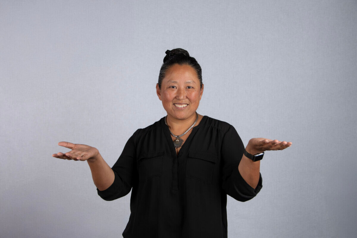 A person wearing a black shirt stands against a plain light gray background, smiling with both hands raised and palms facing upward—capturing the expressive spirit of FACES by Jee Vahn Knight.