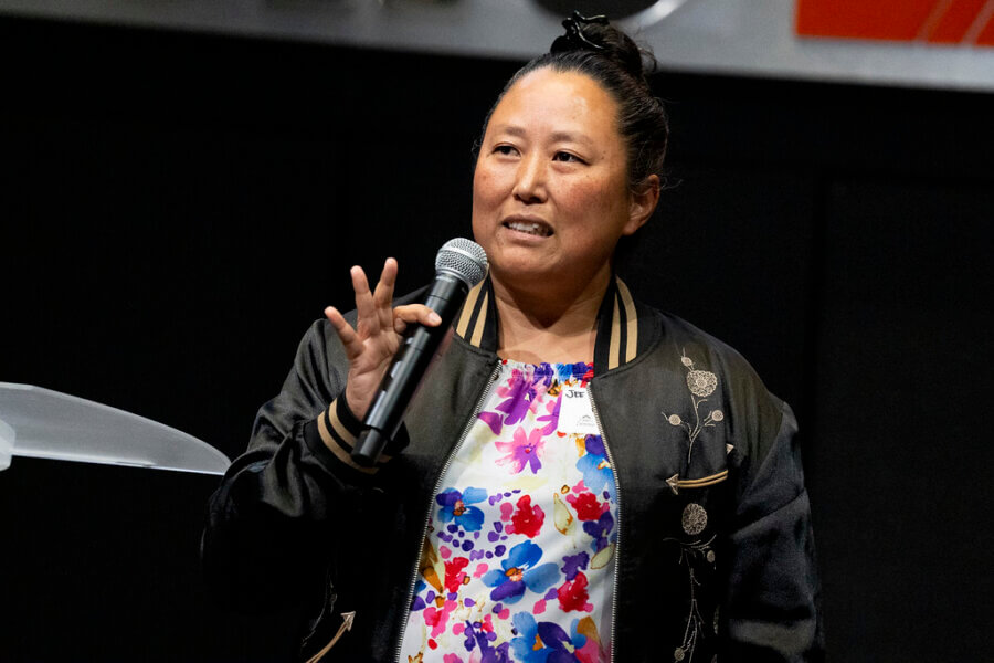 Jee Vahn Knight, wearing a black jacket and floral top, speaks into a microphone while gesturing with their hand on stage at the FACES event.