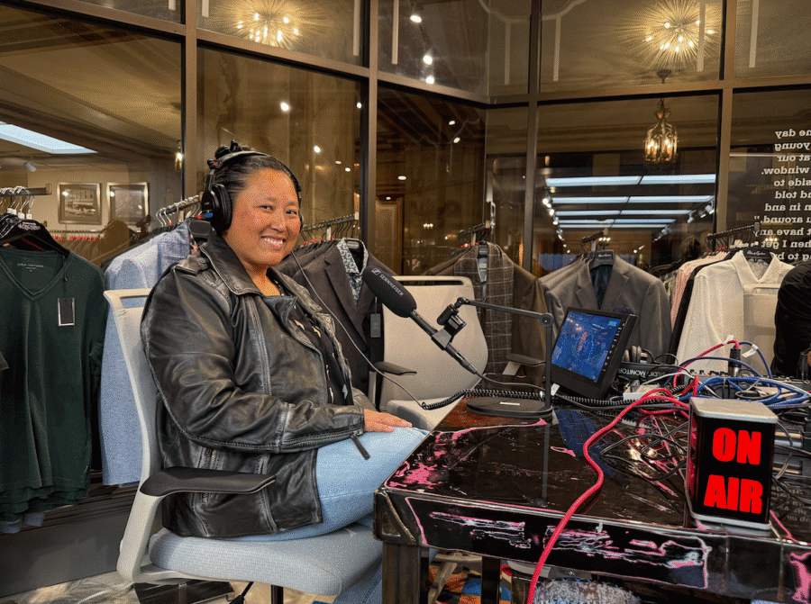 A woman wearing headphones and a black leather jacket sits at a podcast recording setup for Jee Vahn Knight’s FACES in a clothing store, with an "ON AIR" sign lit on the table.