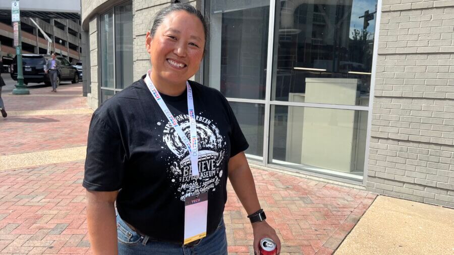 A person wearing a black graphic FACES T-shirt and lanyard stands on a brick sidewalk, smiling and holding a can, with a modern building in the background.