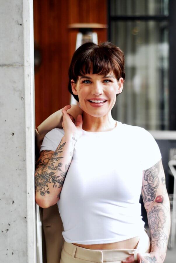 This Master Memphis Tattoo Artist Helps Women Feel Whole Again - 2 Nicolette Tomblyn, featured in FACES, stands outdoors with short dark hair and tattoos on both arms, smiling in a white t-shirt and holding a beige jacket over her shoulder.