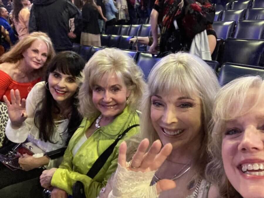 Five women sitting in a row of seats indoors, smiling and waving at the camera; other people are visible in the background.