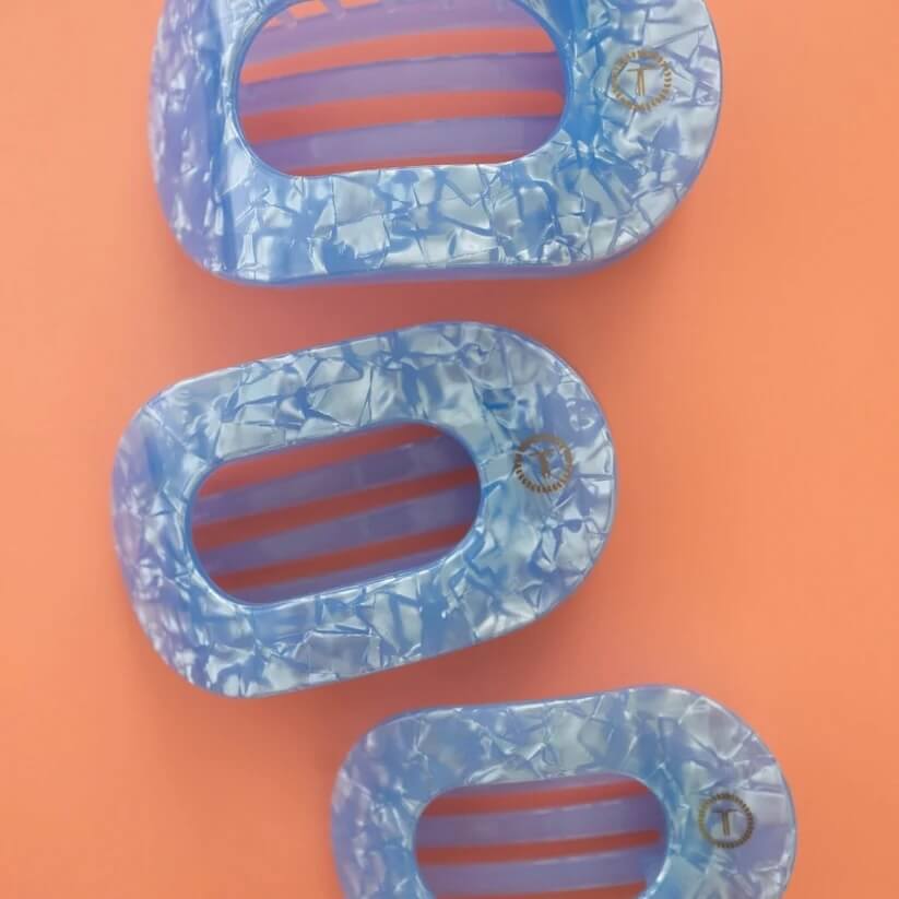 15 Memphis FINDS to Welcome Spring - 8 Three blue, marbled plastic hair claws of different sizes are arranged on a solid orange background—a vibrant look inspired by Memphis finds this February.