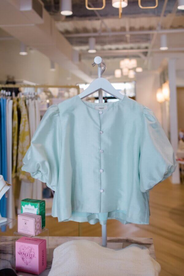 15 Memphis FINDS to Welcome Spring - 2 A light blue, short-sleeve blouse with puffed sleeves and front buttons hangs on a white hanger in a brightly lit clothing store—one of our top Memphis finds this February.