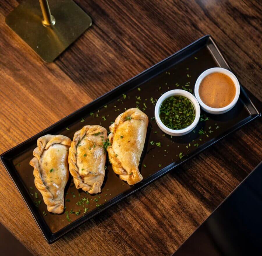 5 New Memphis Restaurants to Try This Month (+ What's Coming Soon!) - 15 Three baked empanadas are served on a rectangular black plate with two small bowls of green herbs and reddish dipping sauce, on a wooden table.