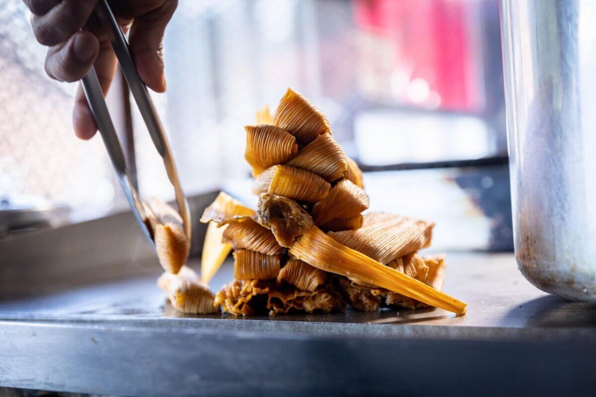 A hand using tongs to pick up pieces of food with a stack of corn husks arranged on a metal surface in the foreground, capturing the essence of Mississippi soul food and its rich food legacy.