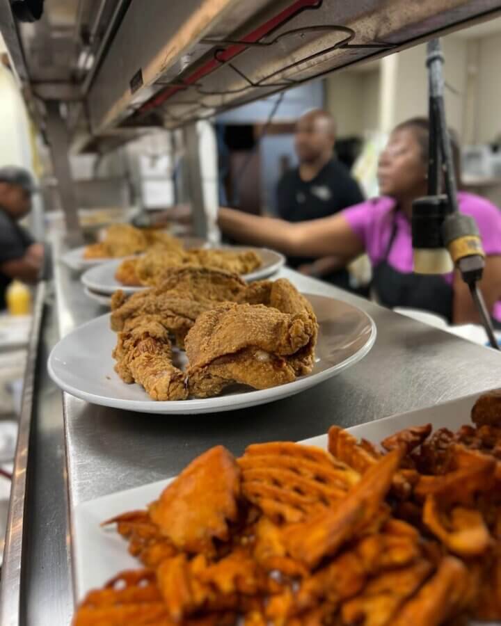 Plates of Soul Food classics like fried chicken and sweet potato waffle fries are lined up on a metal counter, celebrating Mississippi’s legacy, with two people blurred in the background.