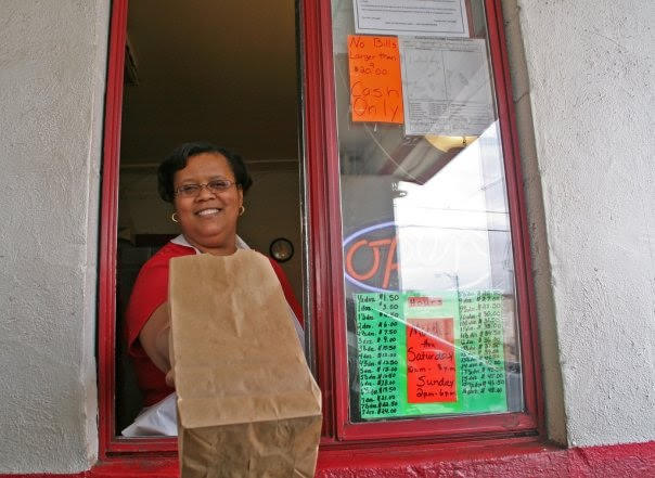 A woman wearing glasses hands a brown paper bag through a takeout window with menu and "cash only" signs visible, serving up Mississippi soul food.