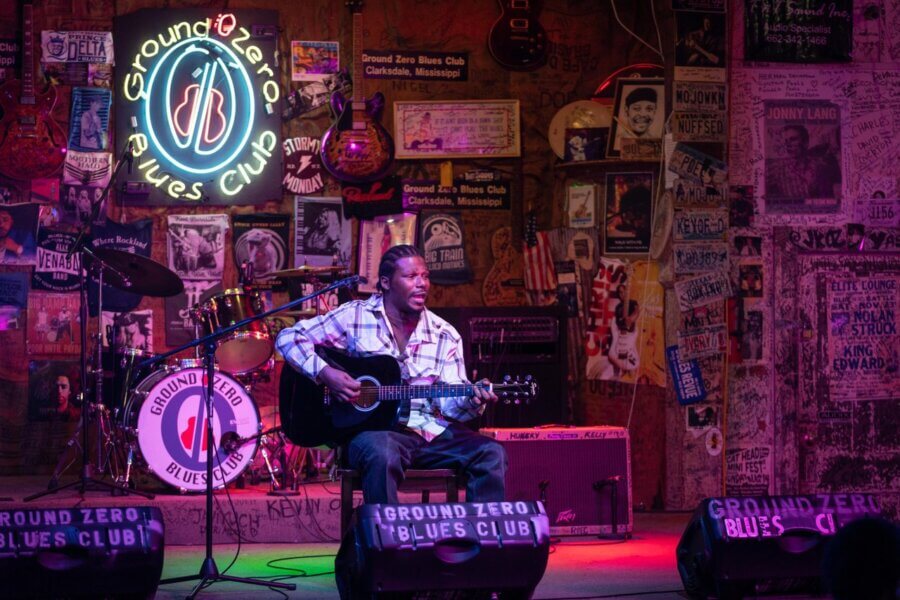 A musician plays an acoustic guitar on stage at Ground Zero Blues Club in Mississippi, surrounded by posters, neon signs, and musical equipment.