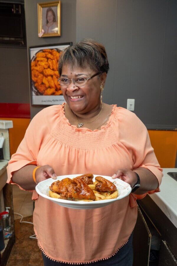 A woman in a peach blouse smiles while holding a plate of sauced chicken wings—classic soul food—in her kitchen. A framed photo of chicken wings hangs on the wall behind her, adding a touch of Mississippi charm to the scene.