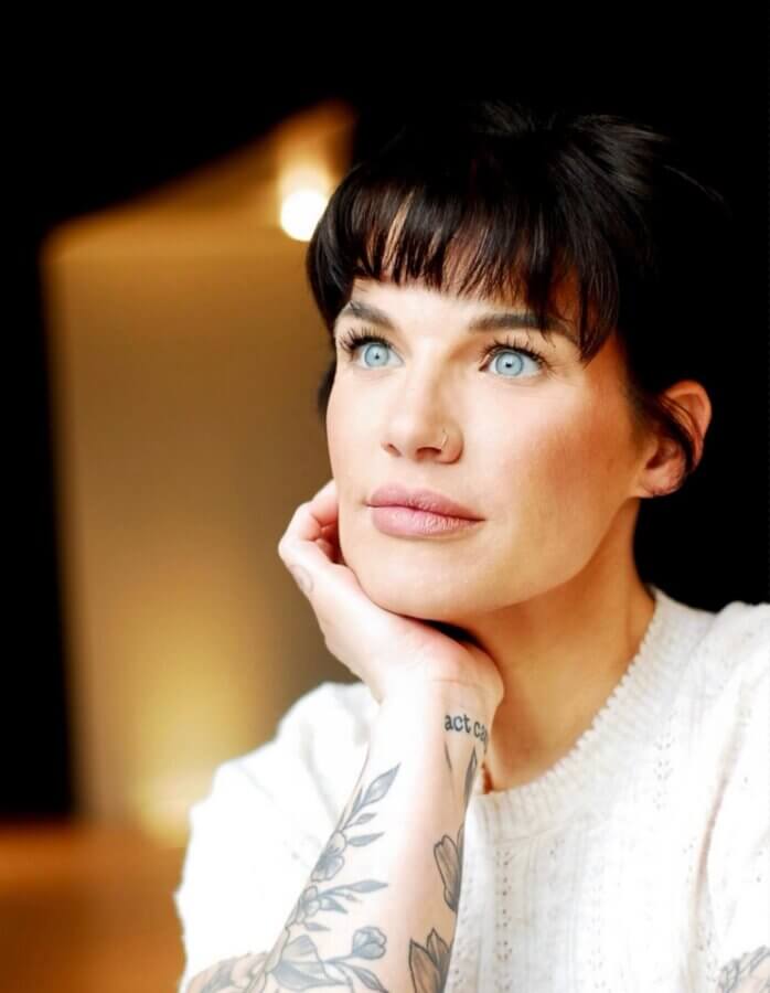 This Master Memphis Tattoo Artist Helps Women Feel Whole Again - 5 A woman with blue eyes and dark hair rests her chin on her hand, wearing a white sweater. Her forearm features a floral tattoo and partial text tattoo. The background is softly blurred, highlighting her unique beauty in the FACES of Nicolette Tomblyn.
