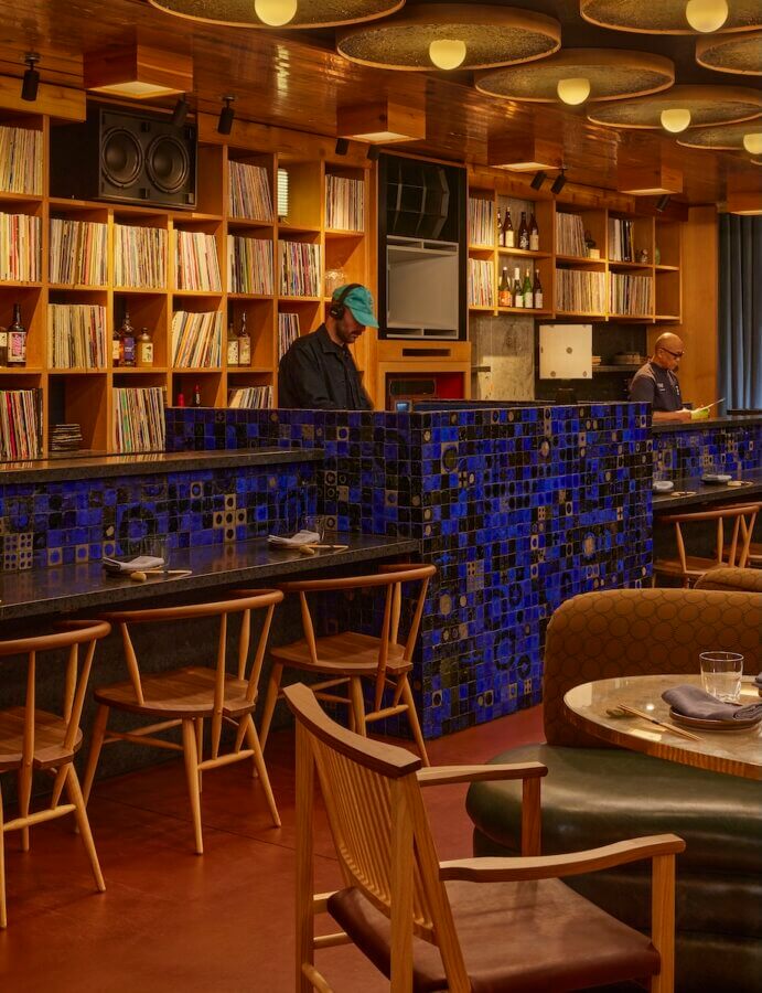 The Rise of Nashville's Vinyl Lounges: 3 to Visit Now + 1 Coming Soon - 4 A restaurant interior with wooden shelves holding vinyl records, a blue-tiled partition, tables and chairs, and two people behind the counter creates a cozy vinyl lounge vibe.