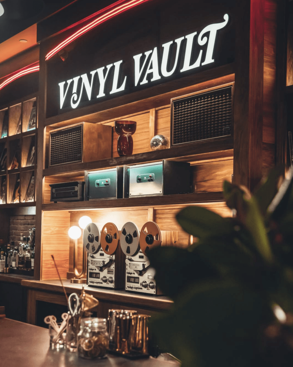 Bar area with “Vinyl Vault” sign, wood shelves holding speakers, tape recorders, and retro decor evoke the charm of a vinyl lounge, with jars and cocktail tools neatly arranged on the counter.