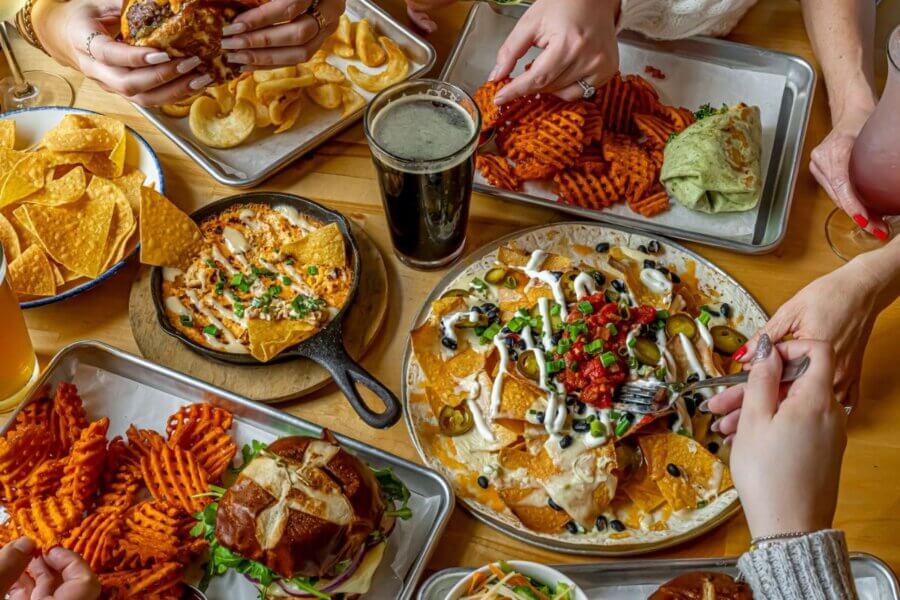 Nashville Steals, Deals & Announcements: March 2026 - 6 A table with nachos, waffle fries, sandwiches, chips with dip, wraps, and drinks; several hands are reaching for food—everyone’s eager to grab a bite of these tasty Nashville steals.
