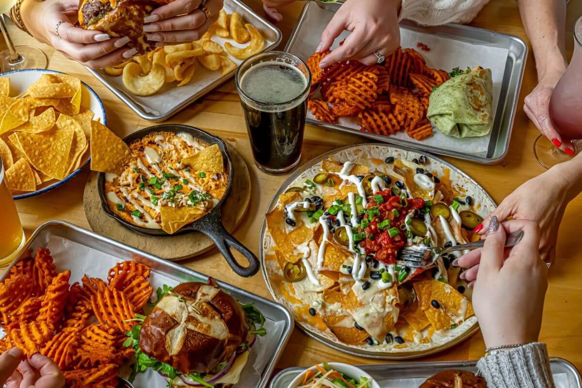 A table with nachos, waffle fries, sandwiches, chips with dip, wraps, and drinks; several hands are reaching for food—everyone’s eager to grab a bite of these tasty Nashville steals.
