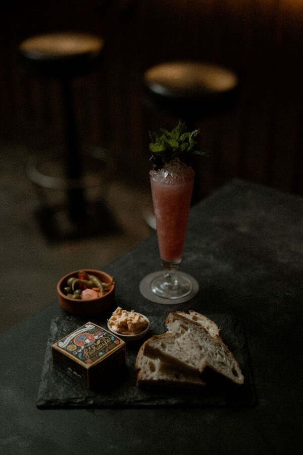 Nashville Steals, Deals & Announcements: March 2026 - 5 A cocktail with mint garnish, a small bowl of mixed olives, a tin container, dip, and bread slices on a dark serving board.